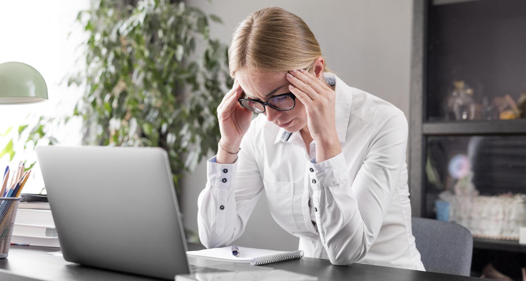 Mulher em escritório com os cotovelos apoiados na mesa e as mãos na cabeça, preocupada com fraudes em documentos.