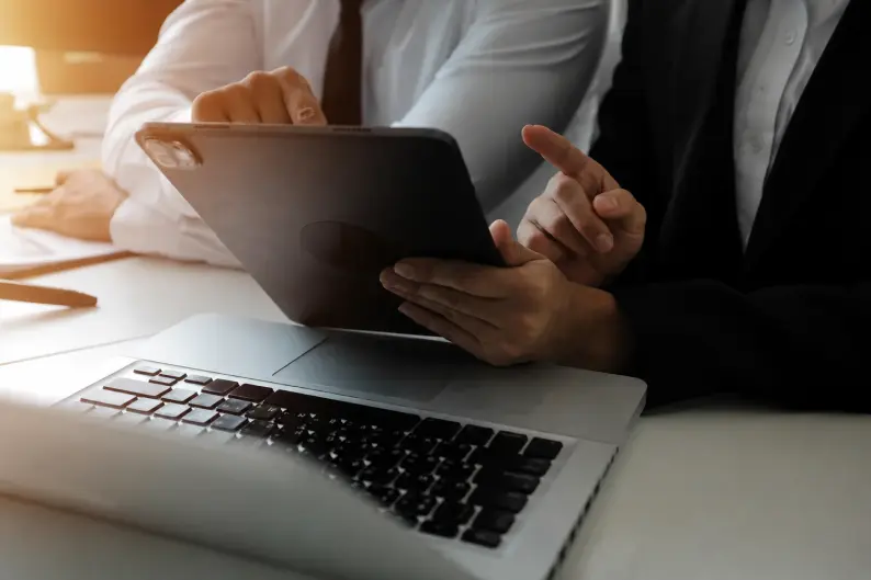 Duas pessoas de roupa social em uma mesa de escritório apontando para a tela de um tablet ao lado de um notebook, analisando um documento digital.