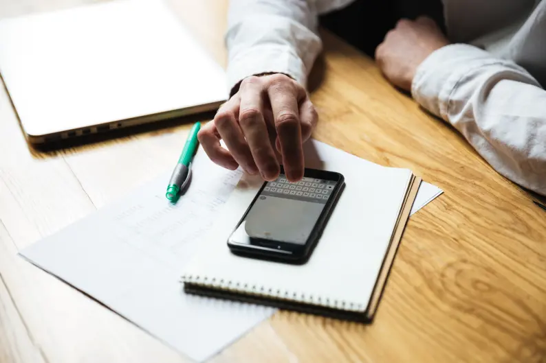 Pessoa em uma mesa de madeira usando um smartphone sobre papéis e um caderno, ao lado de uma caneta e um notebook.